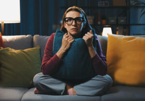 Frightened woman sitting on the couch and watching a horror movie on TV, she is hugging a pillow