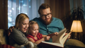 Father, Mother and Little Daughter Reading Children's Book on a Sofa in the Living Room. It's Evening.